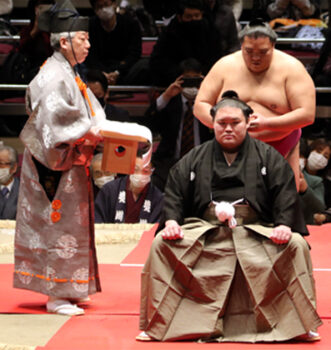 A retiring sumo wrestler in formal montsuki-hakama seated on the dohyo at his danpatsu-shiki retirement ceremony, his oicho-mage topknot still tied