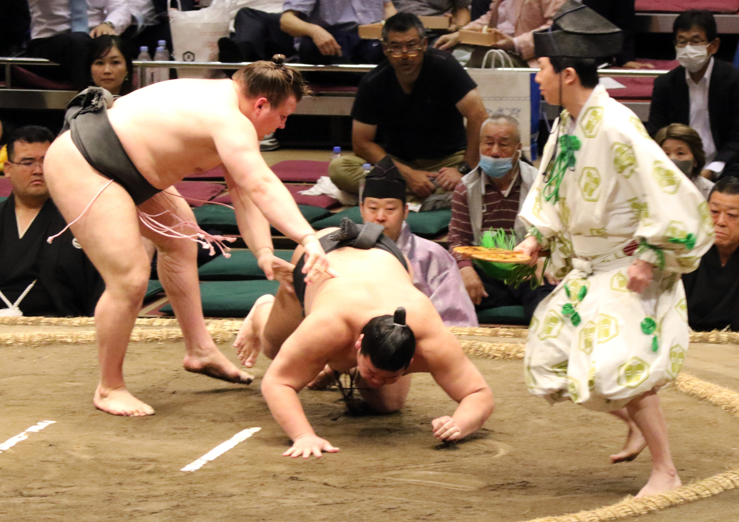 A sumo gyoji in patterned robes moves in as a wrestler goes down on the clay